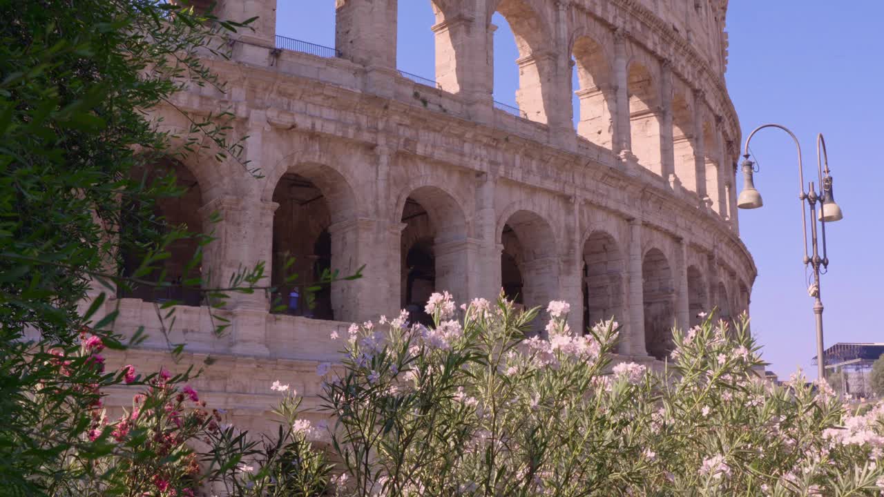 A wide shot highlights the Colosseum's ancient stone arches in Rome, framed by vibrant pink flowers and greenery under a clear blue sky on a sunny day.