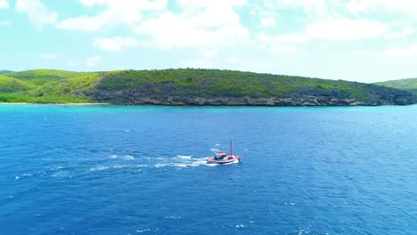 Medium-view-of-small-red-boat-crossing-sea-with-cloud-shadow-on-rocky-shrub-coastline-on-open-ocean