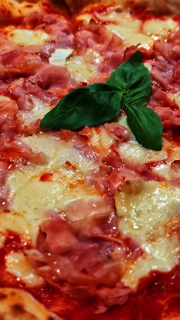 Primo piano verticale di pizza italiana con formaggio fuso, prosciutto, salsa di pomodoro e foglie di basilico fresco, che mostra la consistenza calda e cotta e i ricchi dettagli del cibo da ristorante