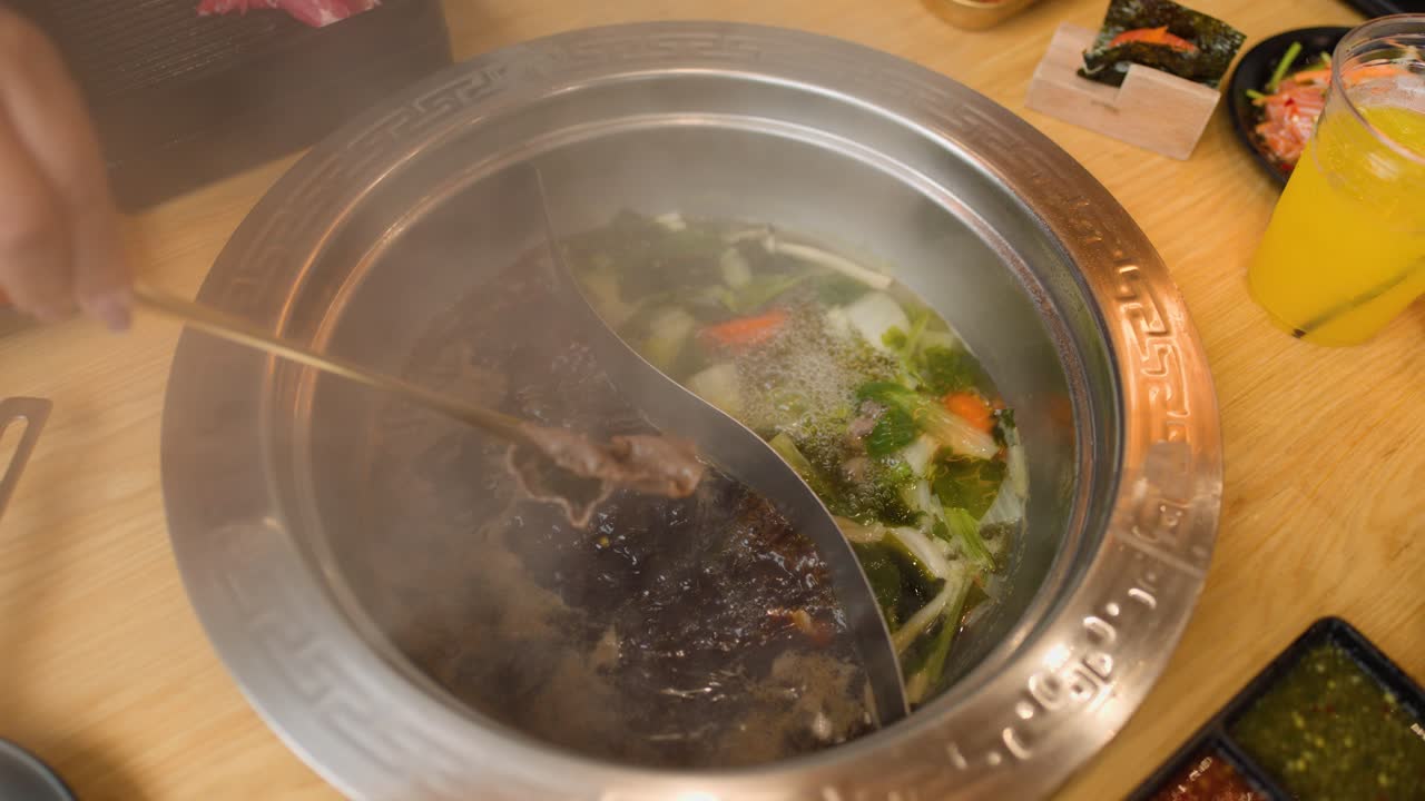 Cooks sliced beef in steaming dual-broth hotpot, surrounded by vegetables, sauces, and side dishes