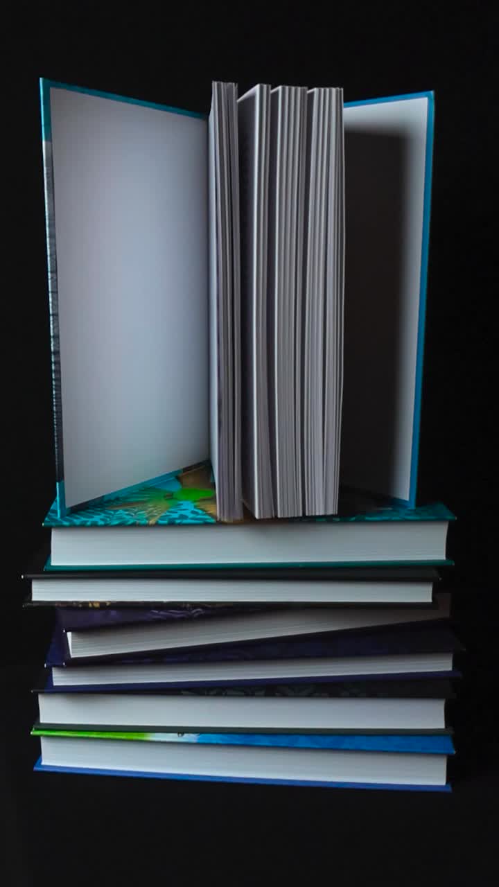 Vertical video of books stacked on top of each other and the one on top is opened. Books have white pages and are hardcovers with colorful covers. Footage spins around them on black studio background.