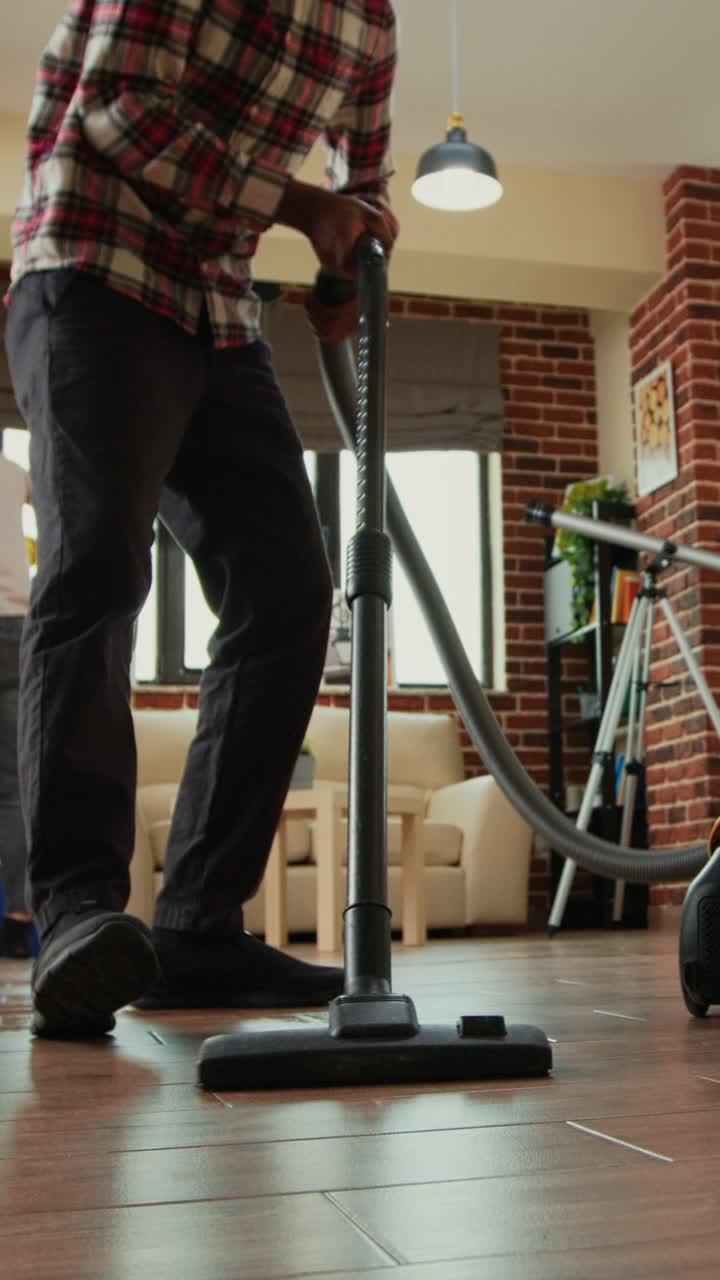Vertical video: African american man vacuuming floors in living room
