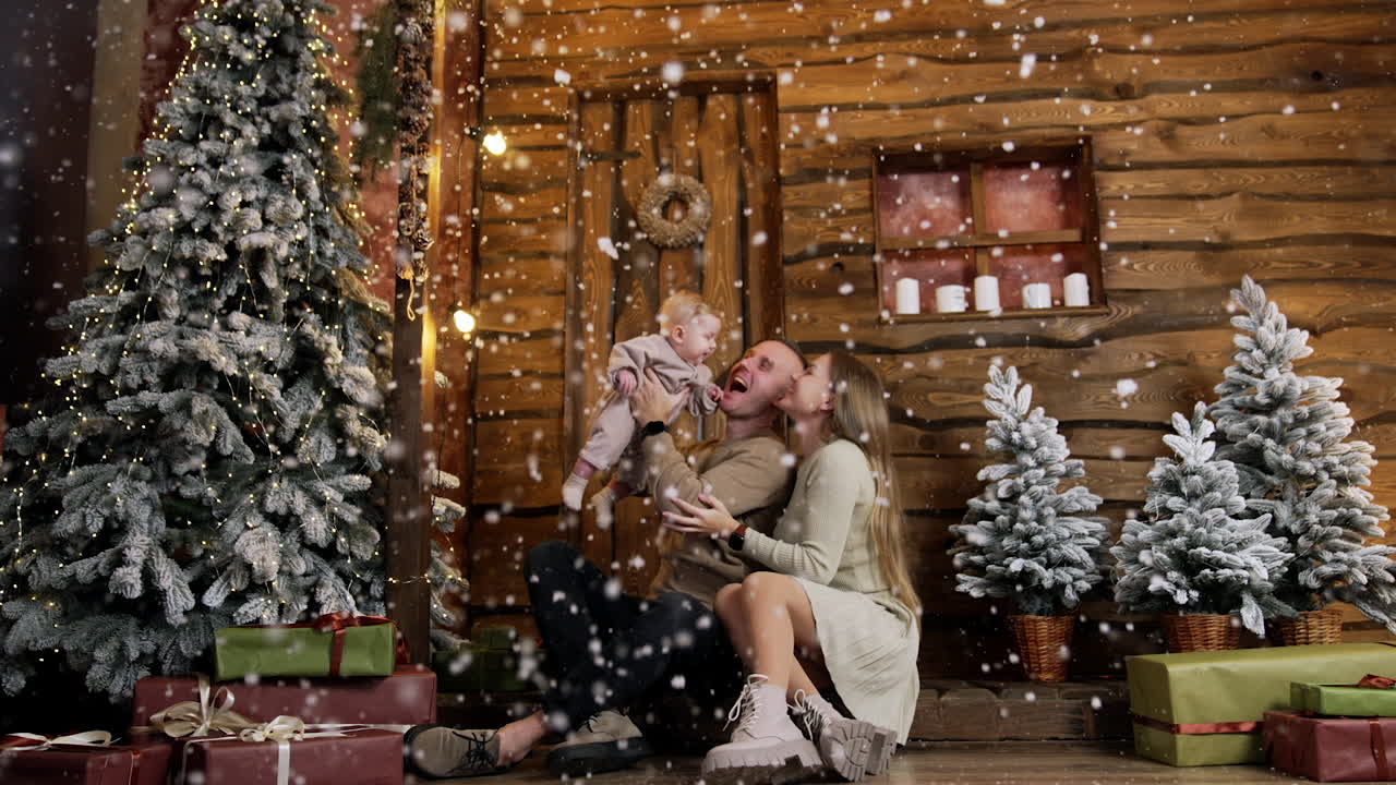 Father raises his adorable baby boy over the head. Happy family enjoys falling snow at the wooden hut at Christmas.
