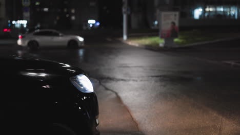 a close-up nighttime view of a black car reversing, with the headlights illuminating the road. another white car can be seen driving in