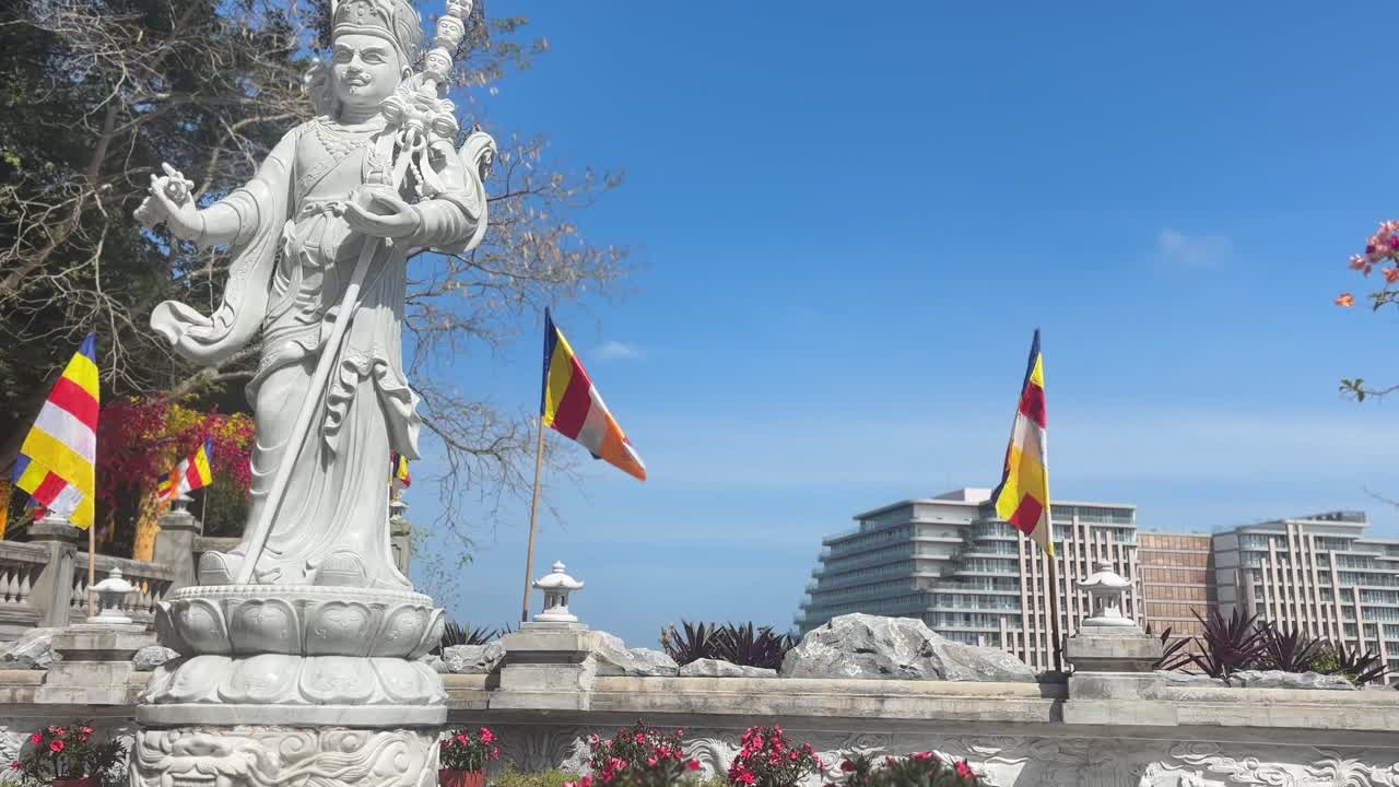Statua buddista nelle Montagne di Marmo Ngu Hanh Son con bandiere colorate, fiori ed edifici della città sullo sfondo, creando una scena culturale all'aperto a Da Nang, Vietnam.
