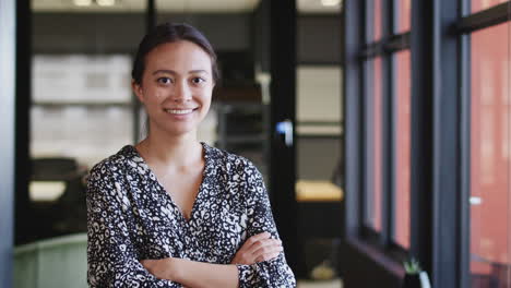 Millennial-Asian-businesswoman-by-window-in-an-office-smiling-to-camera,-waist-up