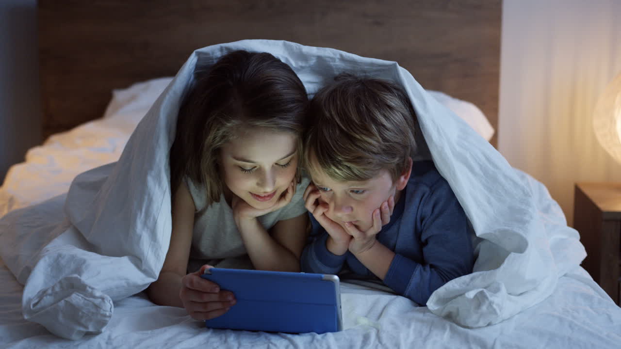 cute small boy and girl lying under the blanket and watching video on the  tablet at night