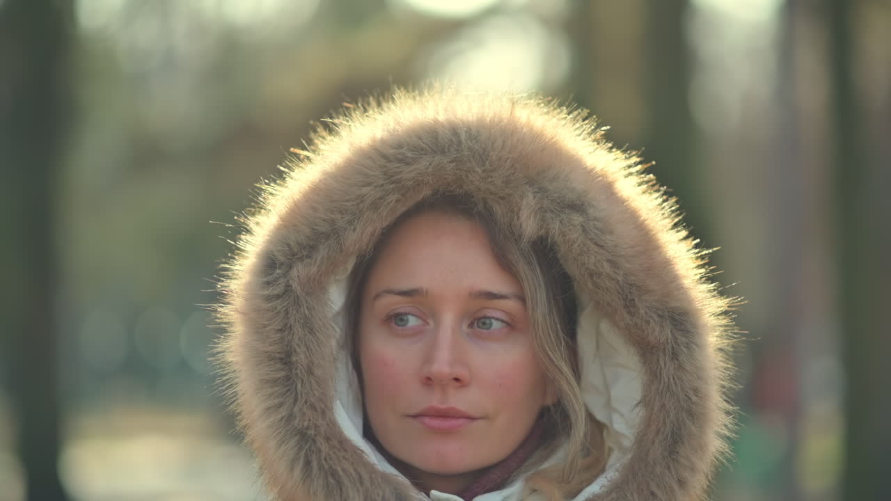 A young woman stands quietly in a snowy park, surrounded by tall trees. She wears a white coat with a fur-lined hood, gazing thoughtfully at her surroundings. The winter sunlight softly shines