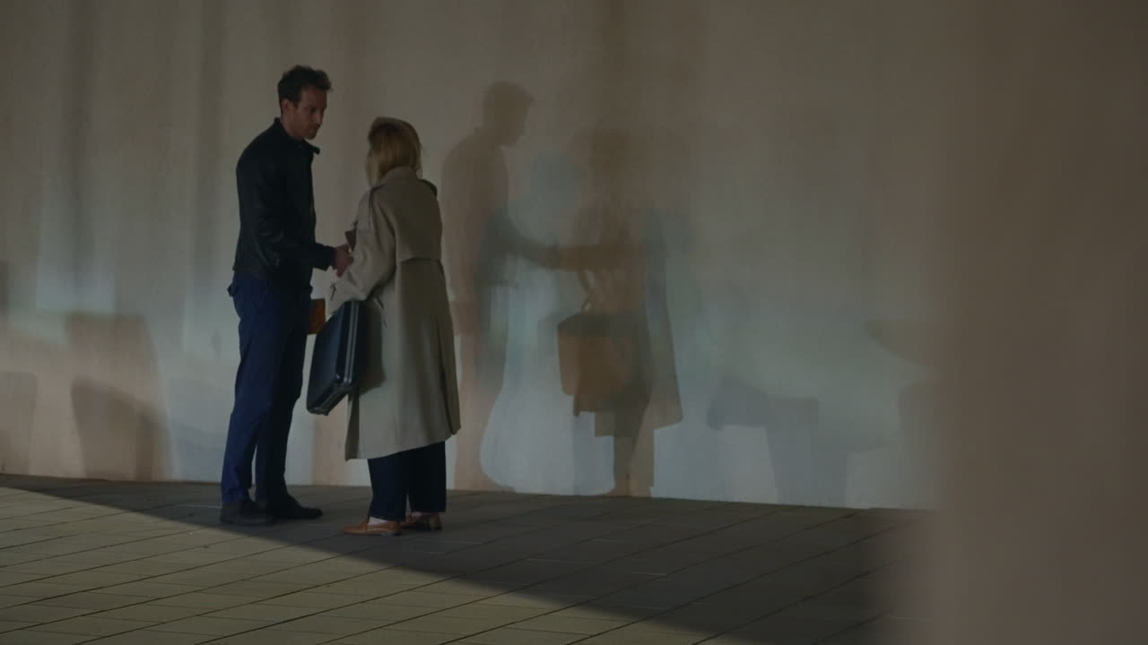 A man and woman secretly exchange a folder and briefcase against a bare wall at night. Their shadows heighten the mood of suspense, secrecy, and covert dealings in an urban setting. Secret agents