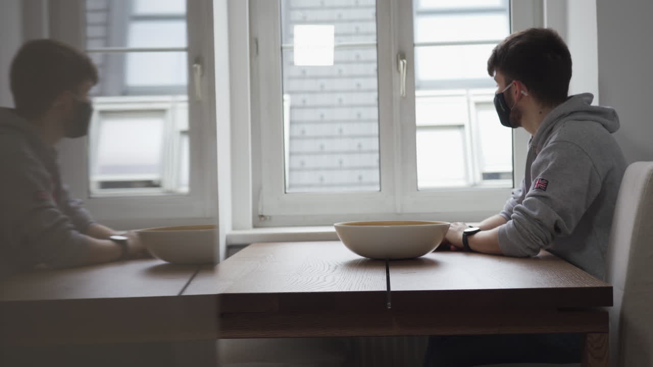 Young man seating with a black mask watching out of the window in times of corona virus.