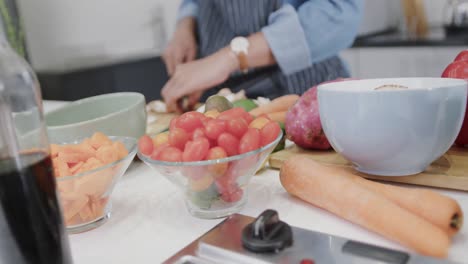 midsection of biracial woman in apron preparing meal, chopping vegetables in kitchen, slow motion