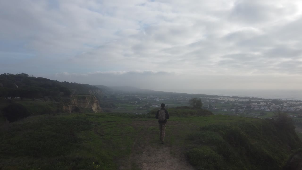 Drone shot following an adventurous man walking towards the end of a cliff with an amazing vista of a coastline.