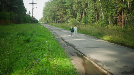 rear view of two children strolling along tarred countryside road with patches of water reflecting nearby trees, surrounded by vibrant green grass and forest under natural daylight