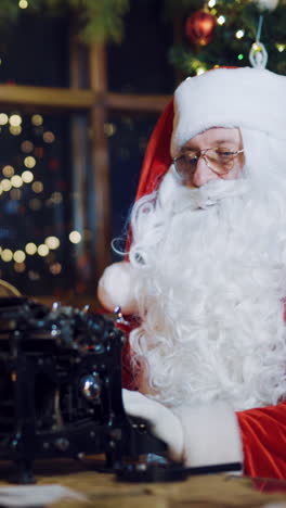 santa claus is sitting and writing letters on the typewriter for children on the background of christmas tree with toys and garlands near window in the room. preparation for new year vertical video