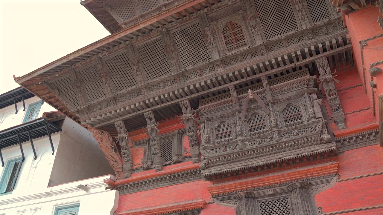 Un plano medio estático contrapicado centrado en un antiguo balcón de madera intrincadamente tallado en una pared de ladrillo rojo de un palacio en la Plaza Durbar, Nepal.