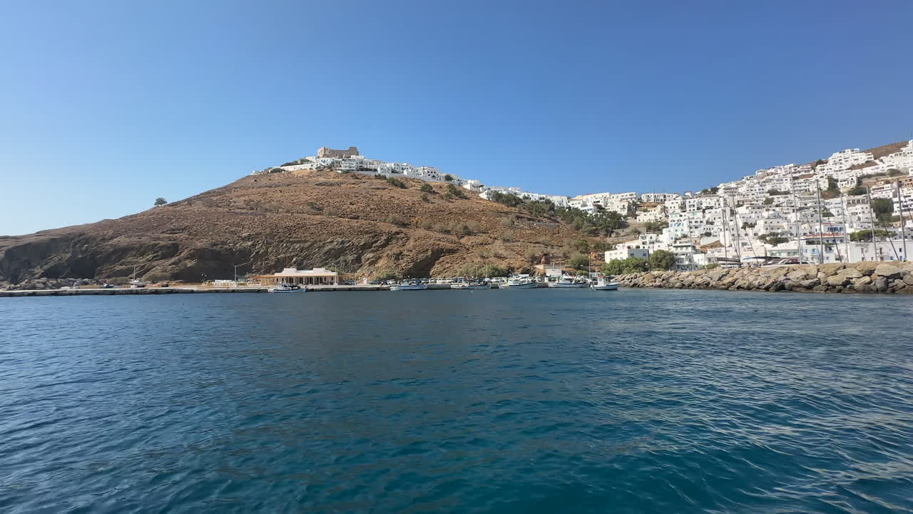 Greece, Astypalea, Castle and Town filmed outside of the harbor wall and from the sea on a sunny day with blue skies. Sea is deep navy blue and very calm.