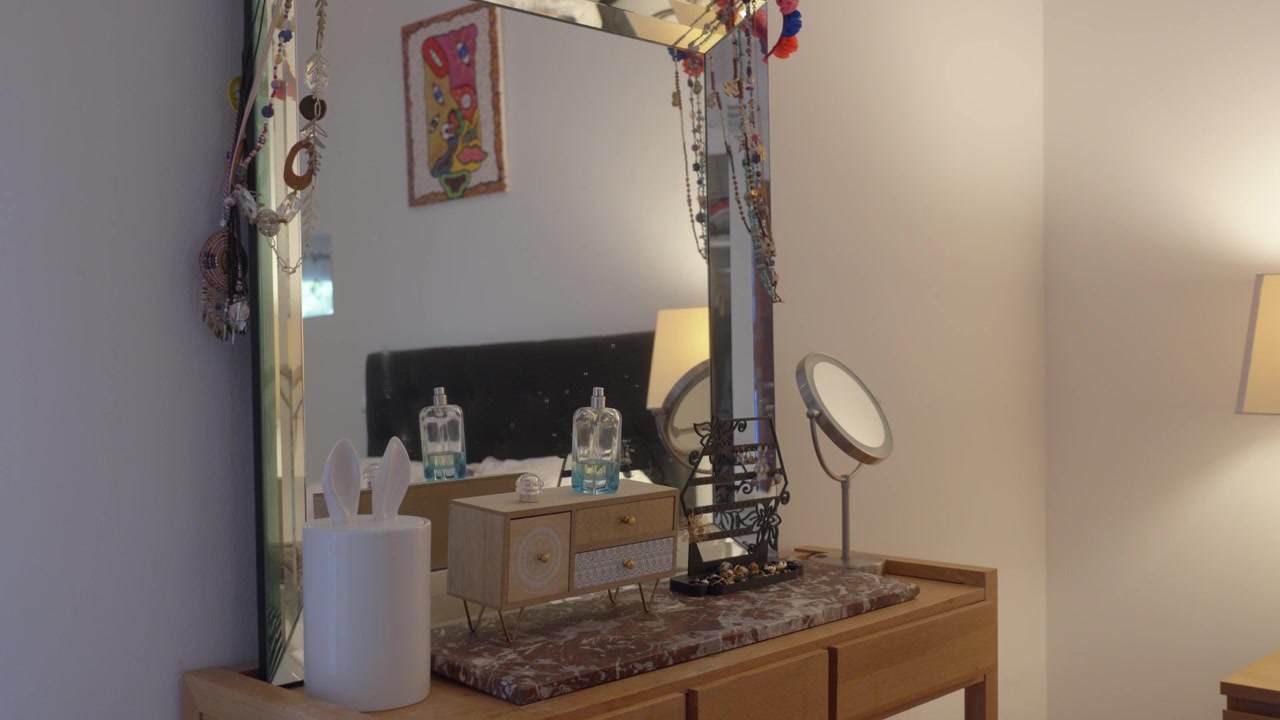 Slow arc shot of a wooden vanity dresser with large mirror, perfume bottles, jewelry organizer, and round vanity mirror in a warmly lit bedroom.