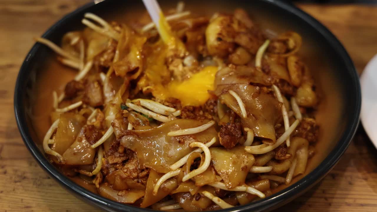 Chopsticks stir a raw egg into a bowl of saucy Singaporean char kway teow noodles with bean sprouts under warm, natural lighting on a wooden table