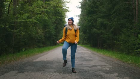 feliz mujer loca viajera bailando y mujer de la calle en suéter amarillo y sombrero con mochila en el camino en el bosque. mujer enérgica con una mochila. el concepto de fronteras abiertas