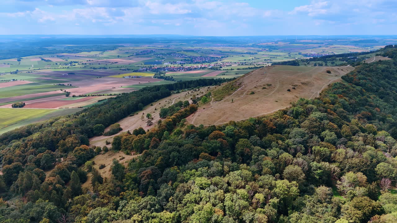 Vista panoramica di colline e campi in una zona rurale con alberi e cielo blu. Colline e campi verdi si estendono nel paesaggio sotto un cielo azzurro.