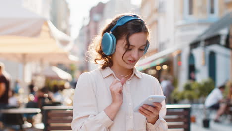 Happy-young-woman-in-wireless-headphones-choosing,-listening-music-dancing-outdoors-city-street