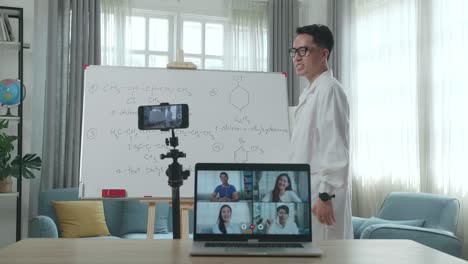 asian male teacher with glasses shoots video by smartphone camera and having video call on laptop while teaching chemist at home