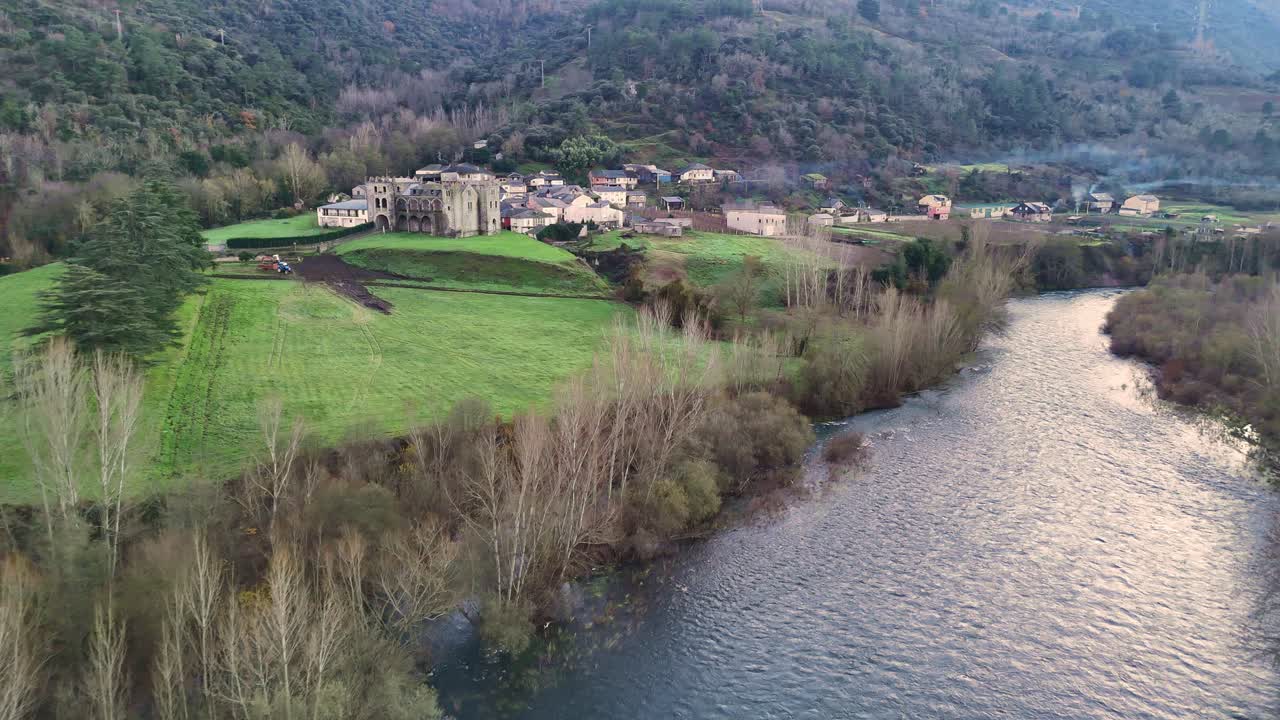 Aerial shot of a winding river leading to a historic stone castle with towers and arches, surrounded by green fields and a village.