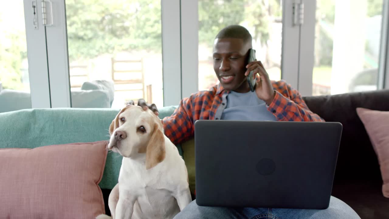 Happy african american man on smartphone and using laptop at home, with his pet dog, slow motion