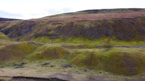 Aerial-drone-shot-flying-towards-some-green-and-brown-moorland-hills-covered-in-heather,-over-a-shallow-stream