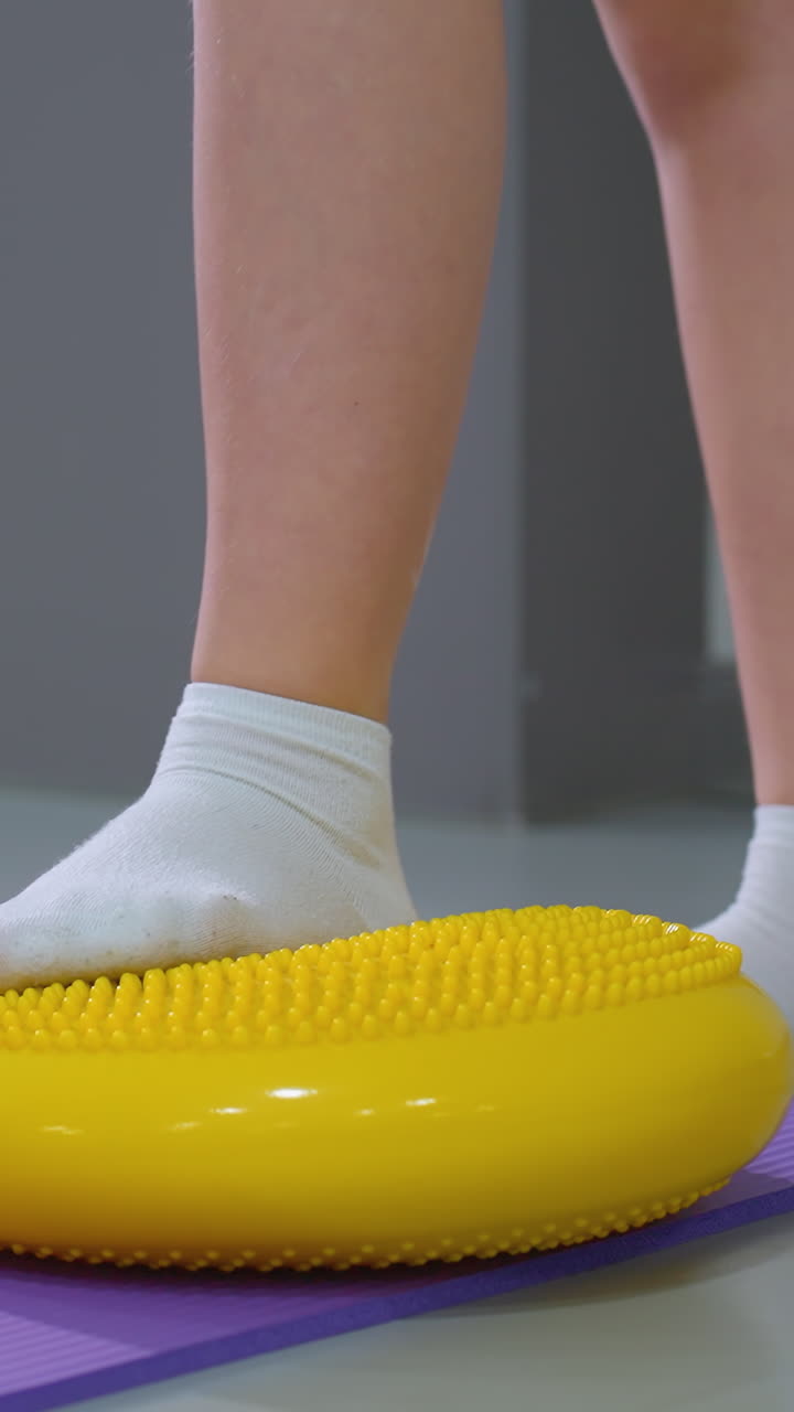 primer plano de un niño pequeño equilibrando en almohadillas de caucho amarillas mientras se arrodilla en una alfombra púrpura, el niño lleva calcetines blancos y pantalones cortos rosados. el fondo presenta equipos de gimnasia