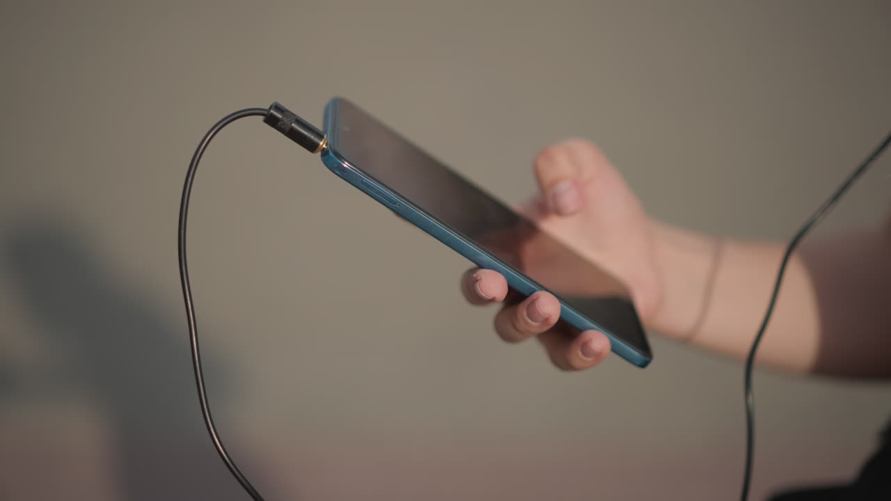 outdoor female plugging phone and headphones on pavement, cropped top and black shorts, checking screen and cable connection, closeup of hands inserting jack and scrolling controls, warm midday light