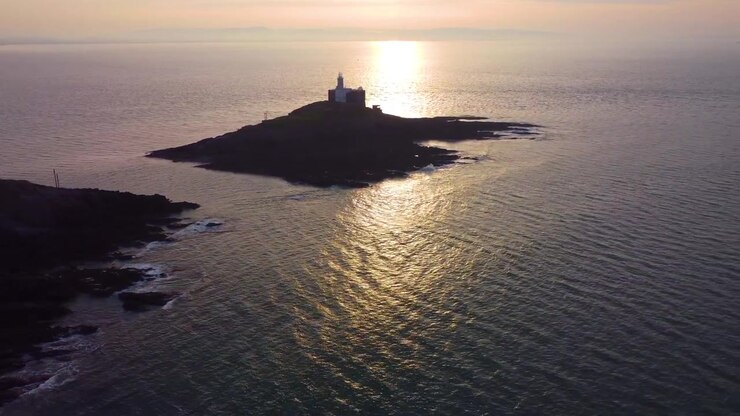 Ampia veduta aerea di Bracelet Bay con il faro di Mumbles all'alba. Riflesso dorato del sole sull'oceano con nuvole dinamiche. Perfetto per il turismo, documentari di viaggio. Galles, Regno Unito. 4K.