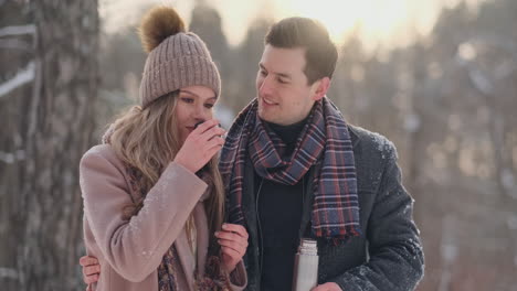 happy couple holding hot tea cups over winter landscape. young couple in love on a winter vacation, standing next to a tree and drinking hot cup of tea