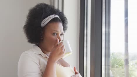 Mujer-Afroamericana-De-Talla-Grande-Mirando-Por-La-Ventana-Y-Sosteniendo-Una-Taza,-Inalterada,-En-Cámara-Lenta