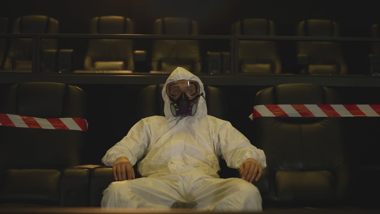 Portrait of a man in white protection costume and respirator sitting alone at the cinema