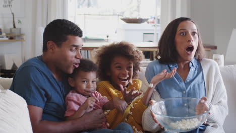 Young-family-sitting-on-sofa-watching-a-movie,-mum-accidentally-spilling-popcorn-in-surprise