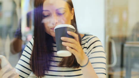 camera focuses on a brunette woman through the window drinking coffee and texting on the smartphone while she is sitting at a table in a cafe