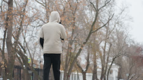 back view of young adult in winter jacket walking and talking on phone with blurred background of dry trees, building, and unclear structures in urban environment on a cold day