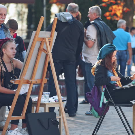 young artists painting on easels. artists drawing pictures on the city street with crowds of people standing nearby.