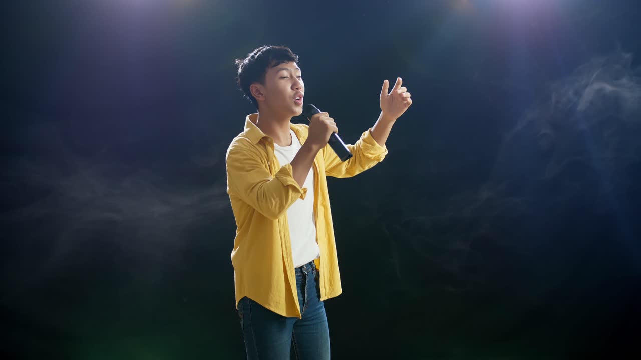 마이크를 들고 있는 젊은 아시아 소년이 색 연기가 있는 검은색 배경에서 을 치고 있다.