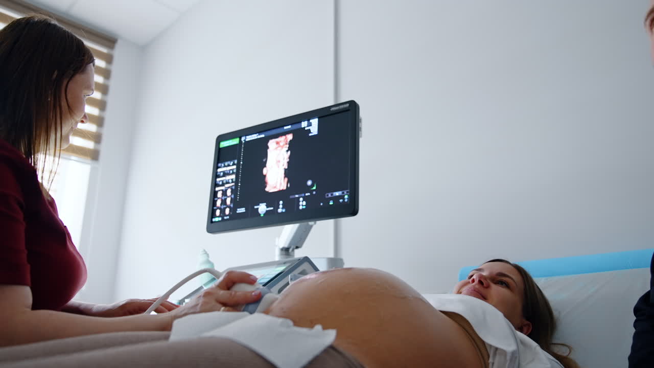 Woman on the last stages of pregnancy is having ultrasonic diagnostics. Expecting woman smiles looking at the monitor seeing her baby.