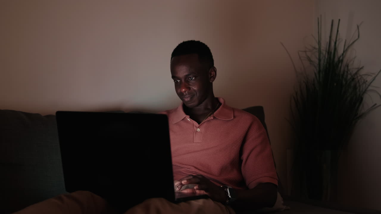Young African Man Working on Laptop on Couch at Night