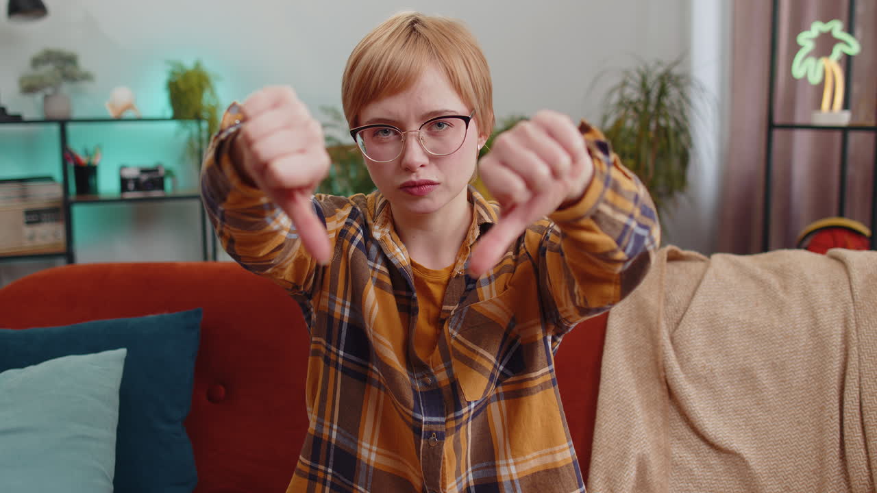 화가 난 젊은 여성은 엄지손가락을 내려놓고, 나쁜 일을 싫어하고, 승인하지 않고, 집에서 불만족스러운 피드백을합니다.