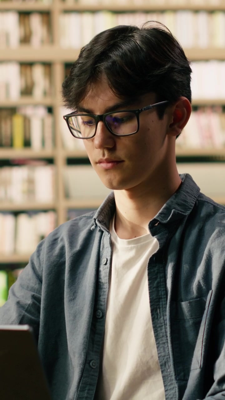 Vertical Video Asian student working on laptop in quiet library with tall bookshelves