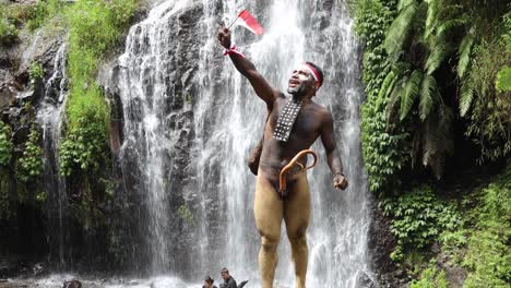 hombre de papua con ropa tradicional de la tribu dani, diadema roja y blanca y brazalete sostiene una pequeña bandera de indonesia y celebra el día de la independencia de indonesia contra el fondo de la cascada