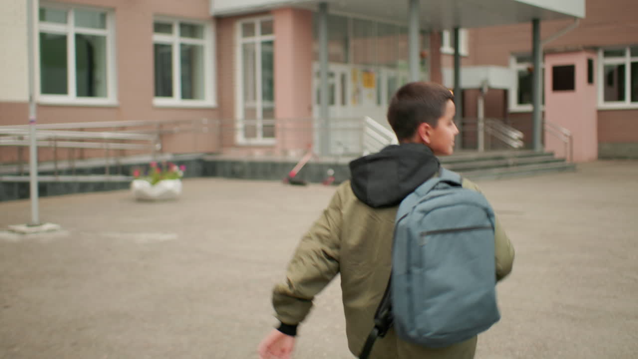 vista trasera de un estudiante con mochila corriendo hacia la entrada de la escuela, marcas del patio visibles en el suelo, exterior del edificio borroso al fondo