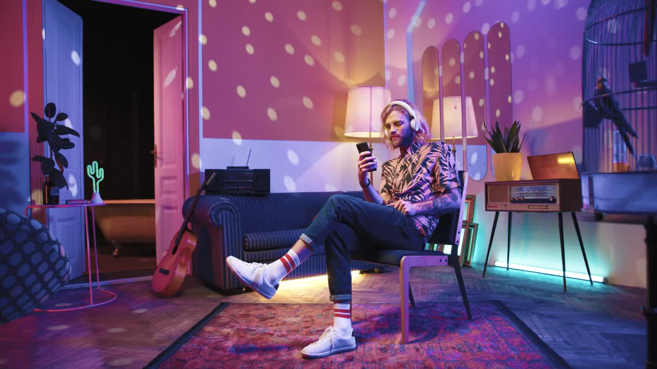 긴 머리에 헤드폰을 끼고 의자에 앉아 집에서 복고풍 파티에서 스마트폰을 사용하는 안경을 쓴 젊은 세련된 백인 남자