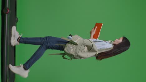 full body of side view asian teen girl student with a backpack holding and reading a book while walking on treadmill in the green screen background studio