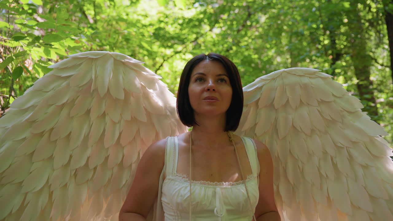 Young woman dressed as angel with large white wings gently taps rhythm on small drum in sunlit forest, surrounded by lush green foliage, expressing creativity