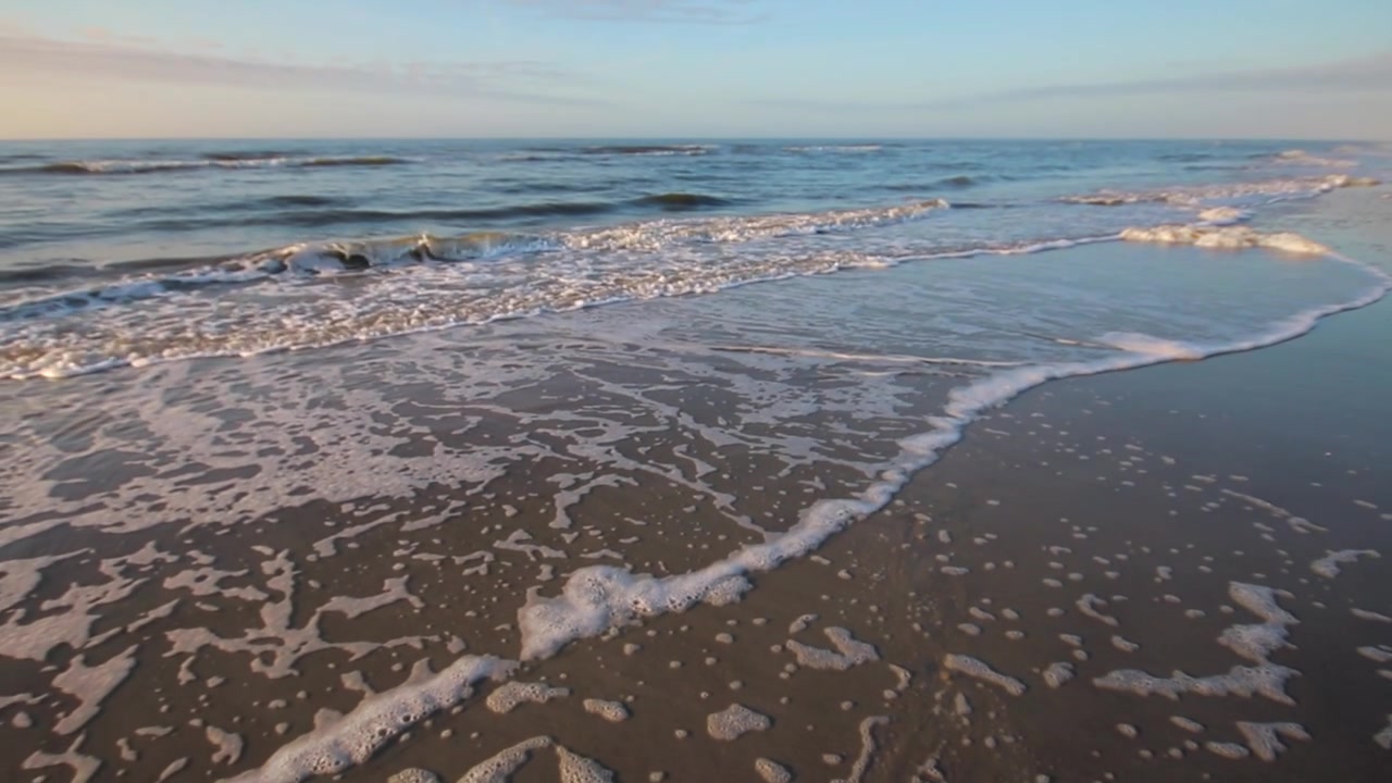 Close-up of ocean waves crashing calmly on beach shore during sunset. Pan right of waves moving on sand on beach by the ocean. Ocean water air bubbles and foam. Slow motion.